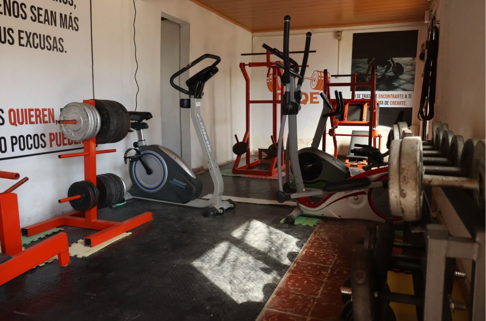 Imagen de las instalaciones de trabajo en la que se ven pesas, maquinas de ejeercicios y bicicletas fijas asi como frases motivavdoras